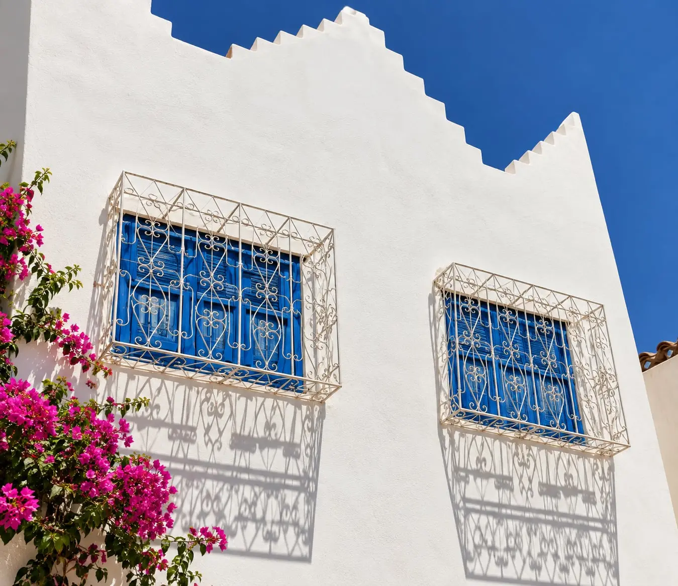 A white Moroccan house with blue shutters and pink bougainvillea